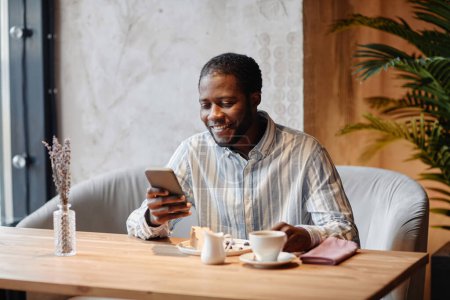 Foto de Joven gerente masculino afroamericano sonriente mirando la pantalla del teléfono inteligente y leyendo noticias en línea mientras pasa tiempo en la cafetería - Imagen libre de derechos