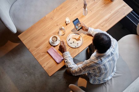 Téléchargez les photos : Au-dessus de l'angle de jeune homme d'affaires avec smartphone prendre des photos de petite cruche avec crème et canapé gris tout en étant assis dans un café confortable - en image libre de droit