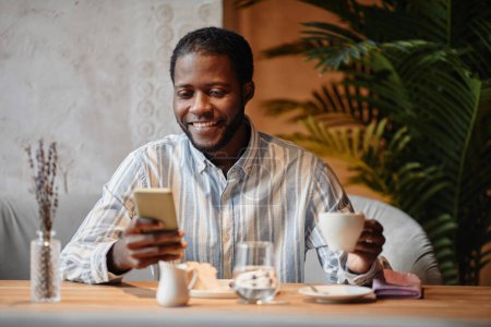 Téléchargez les photos : Jeune entrepreneur afro-américain regardant l'écran du smartphone avec le sourire pendant la communication dans le chat vidéo tout en prenant un café - en image libre de droit