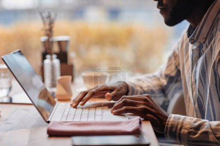 Téléchargez les photos : Mains de jeunes afro-américains pigiste masculin en utilisant un ordinateur portable dans un café tout en étant assis à table et en regardant à travers les données en ligne à l'écran - en image libre de droit