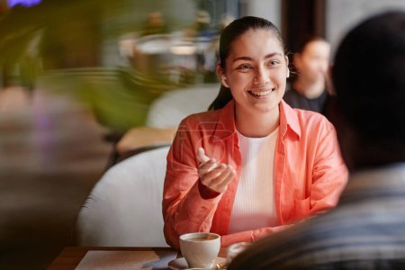 Foto de Joven mujer sonriente con auriculares hablando con el chico afroamericano mientras está sentado frente a él por la mesa en la pausa del café en la cafetería - Imagen libre de derechos