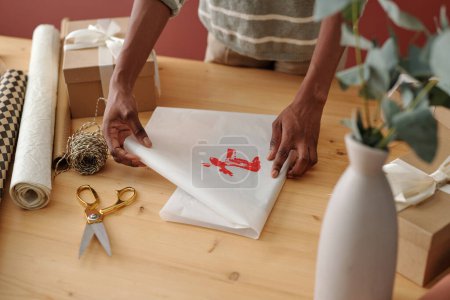 Foto de Chica irreconocible envolviendo regalo en papel blanco con impresión de ciervo rojo hecho a mano mientras prepara regalos de Navidad para amigos - Imagen libre de derechos
