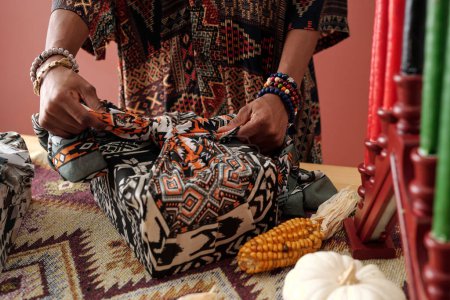 Foto de Mujer negra atando nudo en caja de regalo envuelta en pañuelo decorado con adornos nacionales mientras prepara regalos Kwanzaa - Imagen libre de derechos
