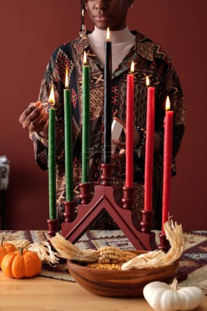 Photo for Young African American woman igniting seven multi-colored candles on candlestick standing on table with pumpkins and ears of corn - Royalty Free Image