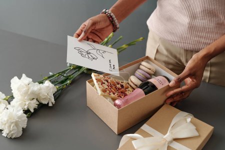 Téléchargez les photos : Femme mettant carte postale à la main avec salutations pour la fête des mères dans une boîte cadeau avec chocolat blanc, macarons et bouteille de champagne - en image libre de droit