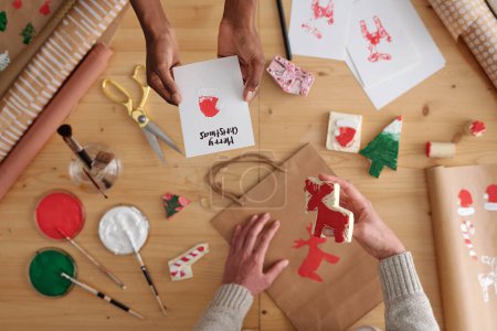Téléchargez les photos : Mains de jeunes femmes interculturelles préparant des cadeaux de Noël tandis que l'une d'elles passe une carte postale de vœux faite à la main à une autre - en image libre de droit