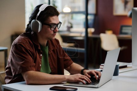 Photo for Side view of young serious designer in headphones typing on laptop keyboard and looking at screen while carrying out new task - Royalty Free Image