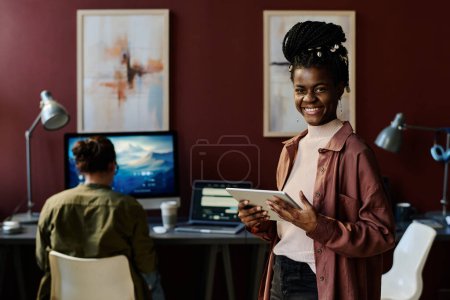 Foto de Feliz joven afroamericana diseñadora femenina con tableta mirando a la cámara mientras está de pie contra su colega masculino en el espacio de coworking - Imagen libre de derechos