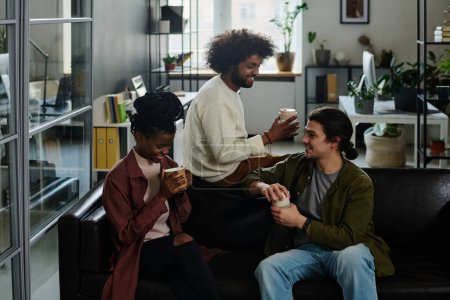 Téléchargez les photos : Trois jeunes designers créatifs prenant un café et bavardant à la pause tout en étant assis sur un canapé en cuir noir dans un espace de coworking - en image libre de droit