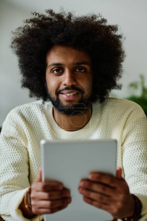 Téléchargez les photos : Jeune homme d'affaires souriant avec tablette regardant la caméra alors qu'il était assis au bureau et faisant défiler les informations en ligne ou regardant la vidéo - en image libre de droit