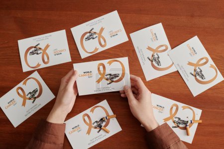 Téléchargez les photos : Au-dessus de l'angle des mains de la jeune femme tenant une carte postale sur une table en bois avec d'autres cartes illustrant le symbole du cancer du poumon et le slogan anti-fumée - en image libre de droit