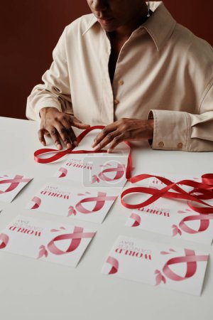 Foto de Joven mujer afroamericana irreconocible con cinta rosa preparando postales antes del día mundial del cáncer mientras está sentada a la mesa - Imagen libre de derechos