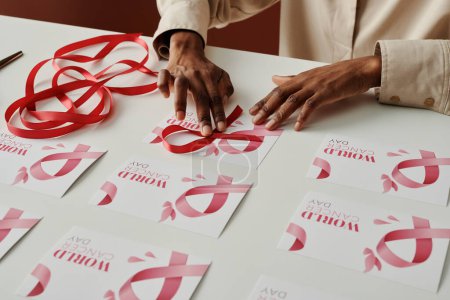 Téléchargez les photos : Mains de jeune femme noire méconnaissable mettre un ruban rose sur la carte postale de la journée mondiale du cancer tout en se préparant pour la campagne de prévention - en image libre de droit