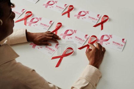 Foto de Primer plano de la chica afroamericana sentada junto al escritorio con un grupo de postales del día mundial del cáncer y poniéndoles cintas rosas - Imagen libre de derechos