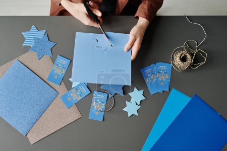 Téléchargez les photos : Au-dessus de l'angle des mains d'une femme méconnaissable découpant les étoiles bleues de David assis sur le bureau avec des cartes postales, du papier et des fils - en image libre de droit