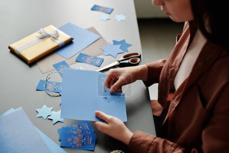 Téléchargez les photos : Jeune femme assise à côté du bureau et préparant à la main Hanoukka cadeaux, cartes postales et symboles avant la célébration de la fête religieuse - en image libre de droit
