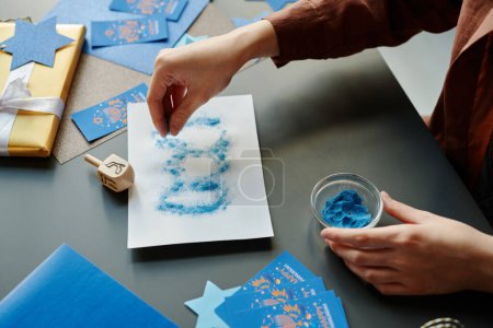 Téléchargez les photos : Main de jeune femme méconnaissable saupoudrant de peinture en poudre bleue sur une feuille de papier avec des contours de dreidel tout en préparant une carte postale créative - en image libre de droit