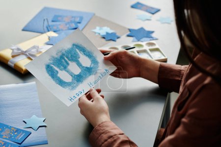 Téléchargez les photos : Fille brune méconnaissable tenant à la main Hanoukka carte postale avec de petits contours de dreidel peint par du sable bleu ou des peintures en poudre - en image libre de droit