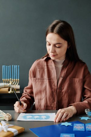 Foto de Joven artesana morena en ropa de casualanotar saludos en Hanukkah postal hecha a mano con fila de contornos dreidel - Imagen libre de derechos