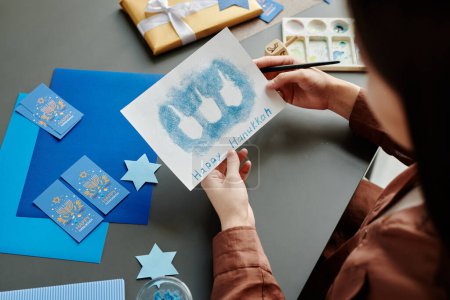 Téléchargez les photos : Angle élevé des mains de la jeune femme tenant Hanoukka carte de vœux avec des contours de petit dreidel sur fond bleu - en image libre de droit