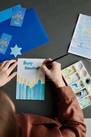 Téléchargez les photos : Angle élevé des mains de la jeune femme écrivant Happy Hanukkah avec pinceau et aquarelle bleue tout en créant de nouvelles cartes postales - en image libre de droit
