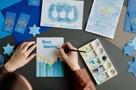 Téléchargez les photos : Mains de jeune femme créative avec pinceau et aquarelle bleue écrivant Happy Hanukkah sur carte de vœux faite à la main - en image libre de droit