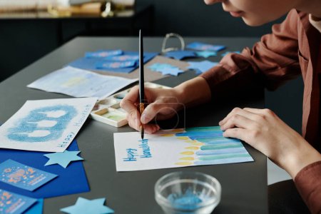 Téléchargez les photos : Jeune artisan avec pinceau se penchant sur le bureau et peignant des bougies allumées sur une nouvelle carte postale créative Hanoukka - en image libre de droit