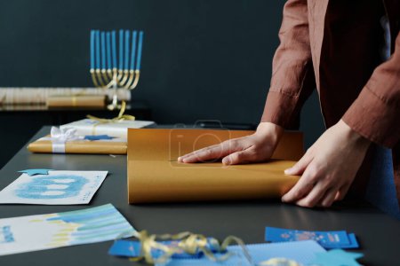 Téléchargez les photos : Mains de fille méconnaissable enveloppant cadeau dans du papier d'emballage beige par bureau avec des cartes postales Hanoukka faites à la main et d'autres articles créatifs - en image libre de droit