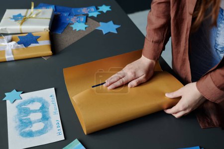 Foto de Manos de una joven irreconocible que envuelve el regalo de Hanukkah en papel beige mientras está de pie junto al escritorio con una postal de Hanukkah hecha a mano - Imagen libre de derechos