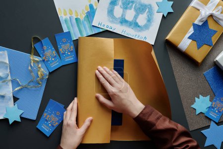 Foto de Vista superior de las manos de una chica creativa irreconocible preparando regalos para Hanukkah y envolviendo uno de ellos en papel beige - Imagen libre de derechos