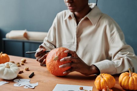 Téléchargez les photos : Plan recadré de jeune femme afro-américaine avec surligneur et citrouille mûre assis par bureau et se préparant pour Halloween - en image libre de droit
