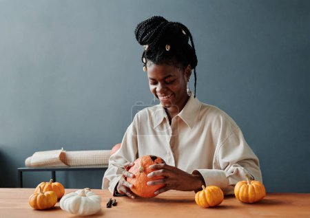 Téléchargez les photos : Jeune femme afro-américaine souriante regardant un visage effrayant sur une citrouille d'Halloween mûre dessinée avec un surligneur noir - en image libre de droit