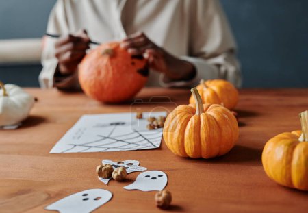 Téléchargez les photos : Groupe de citrouilles orange mûres allongées sur une table avec des fantômes en papier et dessin d'araignées effrayantes contre une jeune femme méconnaissable - en image libre de droit