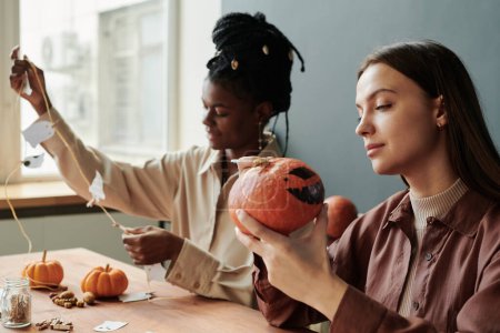 Photo for Young brunette woman looking at spooky Halloween pumpkin in her hands while sitting against female friend creating decorations - Royalty Free Image