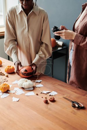Foto de Joven mujer afroamericana poniendo calabazas de Halloween en la mesa junto a otras decoraciones hechas a mano mientras su amiga toma fotos - Imagen libre de derechos