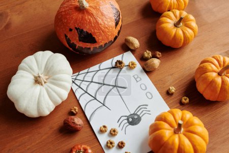 Téléchargez les photos : Carte postale Halloween à la main avec araignée descendant la toile d'araignée allongée sur la table parmi les citrouilles mûres, noix et autres décorations - en image libre de droit