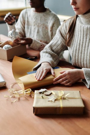 Téléchargez les photos : Jeune femme en pull en laine blanche assise à table devant la caméra et enveloppant Noël présent dans du papier artisanal tout en préparant des cadeaux - en image libre de droit