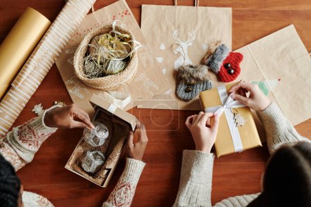 Foto de Vista superior de dos jóvenes mujeres interculturales sentadas a la mesa mientras una de ellas abre una caja de regalo con dos decoraciones de plata para árbol de Navidad - Imagen libre de derechos