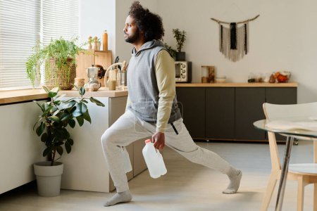 Foto de Joven sosteniendo dos botes de plástico blanco con agua en las manos mientras hace ejercicio físico para estirar las piernas en la cocina - Imagen libre de derechos