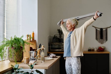 Téléchargez les photos : Femme âgée active en vêtements de sport tenant une serviette au-dessus de la tête entre les bras tendus tout en faisant de l'exercice physique dans la cuisine - en image libre de droit