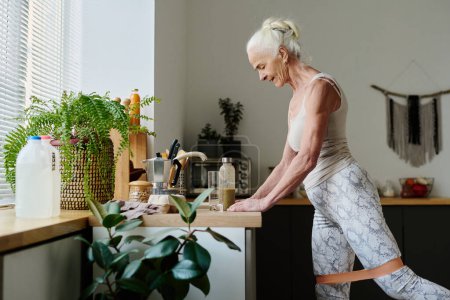 Foto de Mujer anciana activa con banda de resistencia alrededor de las piernas de pie junto al mostrador de la cocina con botella de batido y haciendo ejercicio físico - Imagen libre de derechos