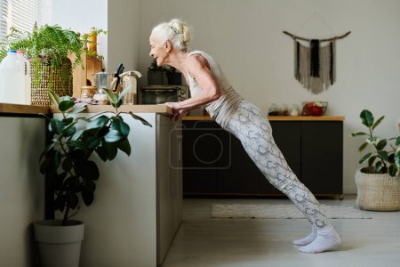 Seitenansicht einer betagten Rentnerin in Aktivkleidung, die beim morgendlichen Training nach dem Schlafen Liegestütze gegen die Küchentheke macht