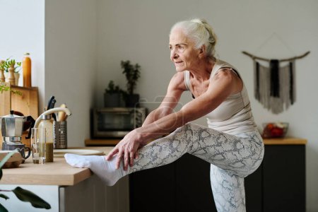 Foto de Mujer mayor activa haciendo esfuerzo mientras estira los brazos sobre la pierna levantada en el mostrador de la cocina y haciendo ejercicios físicos en la mañana - Imagen libre de derechos