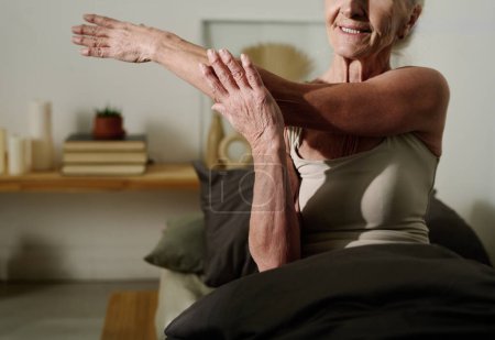 Téléchargez les photos : Plan recadré d'une femme âgée en coton assise sur le lit après le sommeil et étirant le bras tout en faisant de l'exercice physique - en image libre de droit