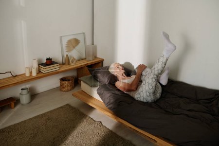 Téléchargez les photos : Boomer féminin actif en vêtements de nuit levant la jambe tendue tout en étant couché sur le lit avec couverture noire et oreiller et faire des exercices physiques - en image libre de droit