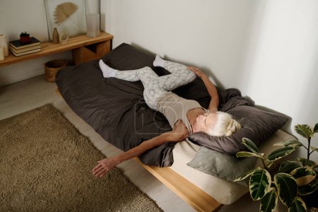 Téléchargez les photos : Angle élevé de la femme active âgée couchée sur le lit après le sommeil le matin et étirant le bras tout en gardant une jambe pliée dans le genou - en image libre de droit