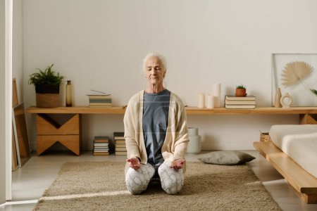 Téléchargez les photos : Femme âgée en tenue décontractée assis sur ses genoux sur le sol de la chambre et gardant les yeux fermés tout en pratiquant l'exercice de yoga - en image libre de droit