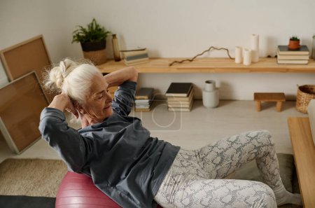 Téléchargez les photos : Femme âgée en vêtements de sport garder sur fitball et pratiquer s'asseoir devant la caméra pendant l'entraînement physique dans l'environnement de la maison - en image libre de droit