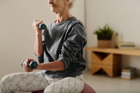 Téléchargez les photos : Femme retraitée en pull gris tenant des haltères assis devant la caméra et pratiquant des exercices pour les muscles des bras après le sommeil - en image libre de droit