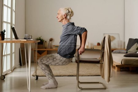 Foto de Vista lateral de la mujer activa envejecida en ropa casual haciendo sentadillas frente a la computadora portátil mientras sostiene la silla y mira la pantalla durante el ejercicio - Imagen libre de derechos